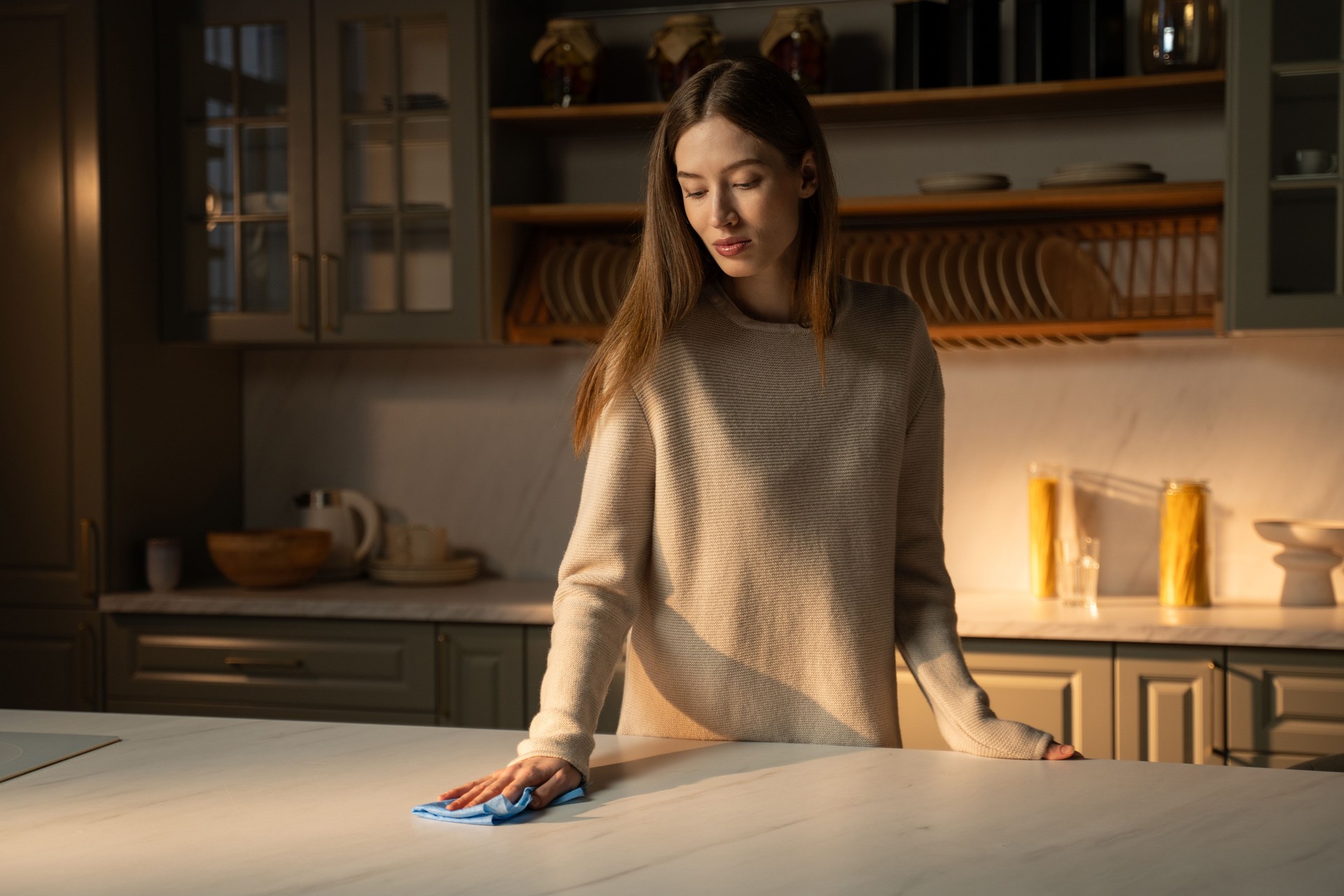 Young Woman in a Sweater Wipes Down a Kitchen Counter in Soft Evening Light